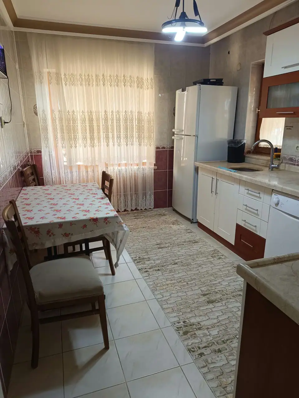 Pencere önünde örtülü yemek masası, buzdolabı ve tezgâh