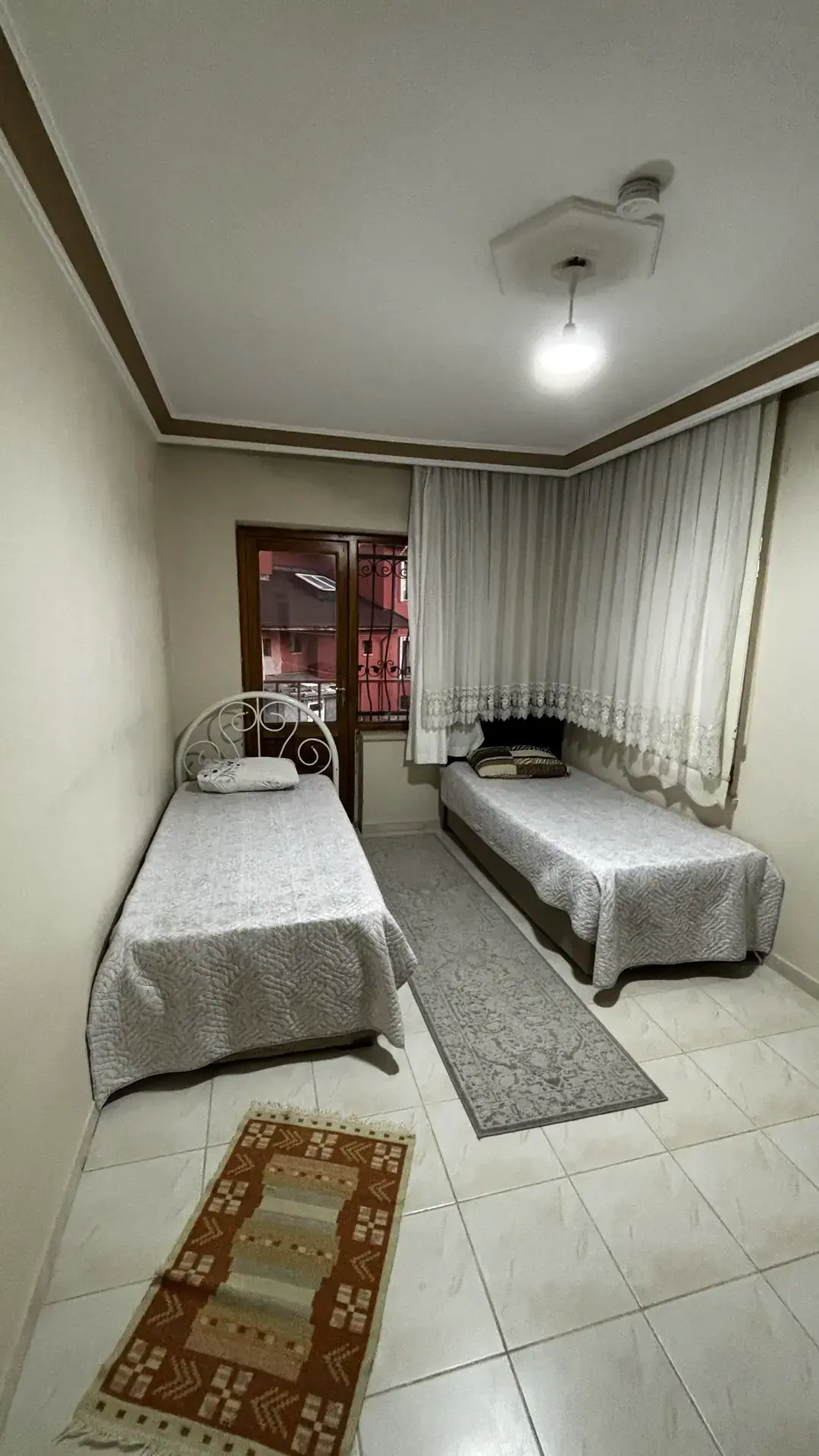 Dantel tüllü pencerenin önünde iki tek kişilik yatak ve el dokuma kilim